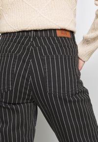 Svartrandiga byxor med vita vertikala linjer, med två bakfickor och en brun läderetikett i midjan. Texturerad tyg.