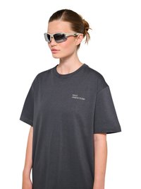 Kobieta ubrana w czarny, luźny T-shirt z napisem „HALO 196819721981” na piersi i białe sportowe okulary przeciwsłoneczne, włosy związane w kucyk.