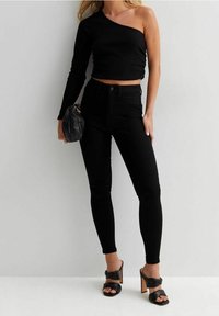 Top corto nero a spalla unica, jeans neri aderenti a vita alta e sandali con tacco neri e cinturini. Tessuto liscio con linee eleganti.
