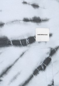 Tela de tie-dye en azul claro y negro con una etiqueta rectangular blanca cosida, que muestra el nombre de la marca "MENNACE" en letras negras.