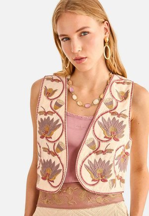 Joven mujer con un chaleco crema con delicados bordados florales en rosa y amarillo sobre una camiseta sin mangas malva, complementada con un collar de cuentas y pendientes de aro.