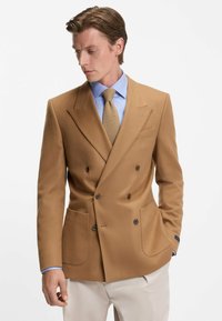 Beige Doppelreiher-Blazer aus glattem Stoff, mit zwei vorderen Taschen und dunklen Knöpfen, getragen über einem hellblauen Hemd und einer Krawatte.