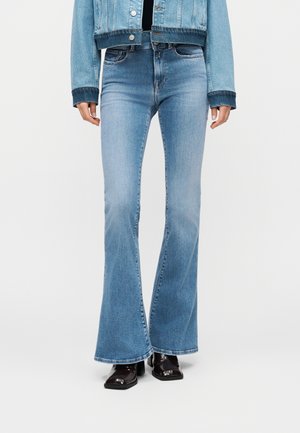 Osoba oblečená v svetlomodrých džínsach s vysokým pásom a zvonovými nohavicami, skrátenej rifľovej bunde a čiernych členkových topánkach s hranatou špičkou na podpätku, stojaca pred jednoduchým pozadím.