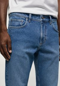 Modré džínové kalhoty s rovnými nohavicemi, s přední kapsou, zapínáním na knoflík a viditelnými švy. Hladká textura.