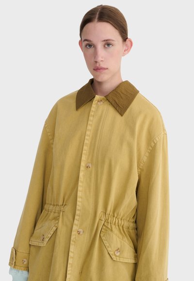 Giovane con pelle chiara e capelli castani raccolti, che indossa una giacca giallo senape con ampio colletto e tasche frontali con bottoni.