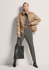 Beige Daunenjacke mit hohem Kragen, schwarze gemusterte Bluse, gemusterte Hose und schwarze Ledertasche mit quadratischem Boden und Griffen.