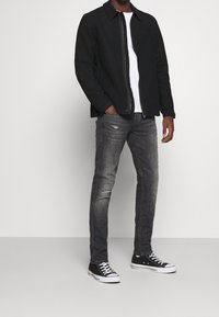 Chaqueta negra, camiseta blanca, vaqueros grises desgastados y zapatillas negras. Diseño sencillo, ajuste casual y roturas notables en los vaqueros.