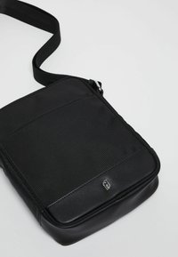 Borsa a tracolla nera realizzata in un tessuto liscio con un pannello frontale testurizzato e un fondo morbido. Presenta un logo semplice e una tracolla regolabile.