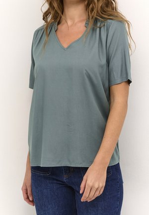 Femme portant un chemisier vert sauge à manches courtes et col en V, assorti à un jean bleu foncé, debout devant un fond blanc.