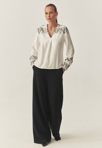 Blouse blanche avec broderie florale noire autour de l'encolure et des manches ; associée à un pantalon large noir et à des chaussures pointues noires.