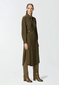 Majica u maslinastoj boji s dugim rukavima, s pojasom u struku, sa bočnim prorezima i glatkom teksturom, u kombinaciji s sjajnim smeškim čizmama do koljena.