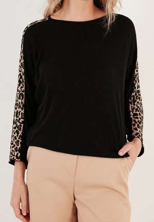 Vrouw draagt een zwarte top met lange mouwen, met beige luipaardprintpanelen op de mouwen en lichtbeige broek, één hand in de zak.