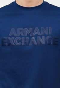Μπλε μπλούζα με στρογγυλή λαιμόκοψη και ανάγλυφα σκουρομπλε γράμματα "ARMANI EXCHANGE" στο στήθος, φορεμένη από ένα άτομο που φαίνεται από τον λαιμό και κάτω.