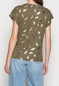 Top de manga corta en verde oliva con patrones de hojas metálicas doradas, corte relajado, tejido texturado y diseño de cuello redondo.
