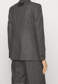 Dopasowana marynarka z ciemnoszarego flanelu z klapami z wcięciem, zapinanymi na guziki mankietami oraz czarną ozdobną metką z tyłu. Czyste linie i struktura.