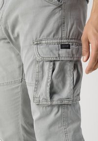 Jambe droite d’un pantalon cargo gris clair avec poche à rabat et une main reposant à côté sur un fond uni.