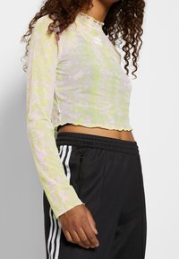 Eine Frau trägt ein langärmliges, gelbes und hell-lila Schlangenmuster-Crop-Top und schwarze Adidas-Trainingshosen mit weißen Streifen und Reißverschlusstaschen.
