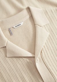 Beige stickad skjorta med krage som har ett strukturerat mönster, knappstängning och subtil ruggning för extra detalj. Etiketten visar "GABBA."