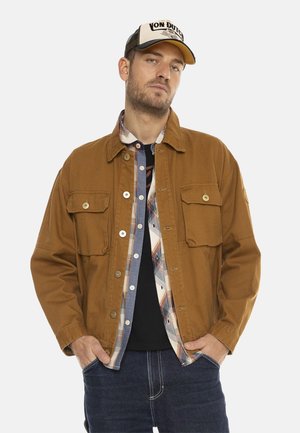 Homme portant une casquette beige, une veste marron par-dessus une chemise à carreaux et un t-shirt noir, debout les mains dans les poches sur un fond blanc.