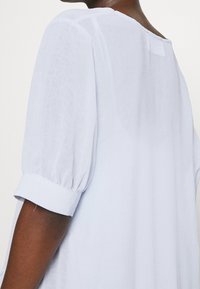 Světle modrá blůza s texturovanou tkaninou, kulatým výstřihem a krátkými nadýchanými rukávy s manžetou. Detaily na zadní straně nejsou viditelné.