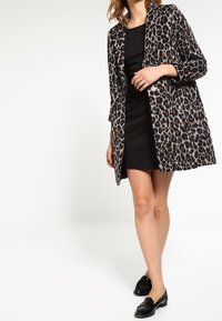 Płaszcz w cętki z długimi rękawami, z przodem zapinanym na guziki i luźnym kroju, noszony na czarnej sukience bez rękawów, w zestawieniu z czarnymi butami.