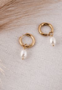 Cercei cerc aurii cu perle albe în formă de picătură, așezați pe o țesătură bej texturată, alături de iarbă uscată decorativă.