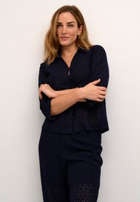 Camicia in pizzo blu navy con chiusura a bottoni sul davanti, vestibilità comoda, maniche a tre quarti e orlo a strati, abbinata a pantaloni in pizzo coordinati.