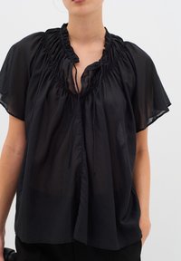 Femme portant un chemisier noir ample et transparent avec un col froncé et des manches courtes, tenant un sac à main noir à son côté.