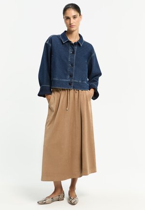 Frau mit dunklem Haar im Dutt, die eine dunkelblaue Jeansjacke, einen beigefarbenen Midirock mit Kordelzug und flache Schuhe mit Schlangenledermuster trägt, steht mit den Händen in den Taschen.