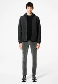 Giacca zip grigia scura con cappuccio nero, indossata sopra una maglietta nera, abbinata a pantaloni slim-fit grigi e sneakers nere. Texture liscia.