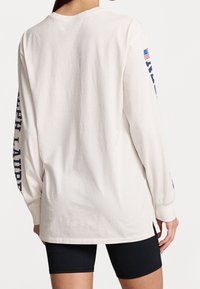 Vit långärmad t-shirt i bomull med blå text på ärmarna och en liten amerikansk flagga som accent.