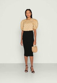 Blusa bege de mangas bufantes com pontos brancos, combinada com uma saia midi preta justa e sandálias pretas de tiras. Segurando uma pequena bolsa de palha.