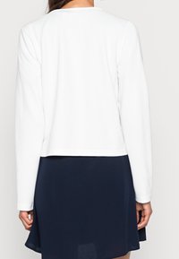 Top bianco a maniche lunghe con collo a giro, realizzato in tessuto testurizzato, indossato sopra una gonna blu scuro. Sono visibili linee pulite e un design cropped.