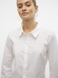 Mladá žena s zlatými kruhovými náušnicemi a bílou košilí s knoflíky na pozadí čistého bílého pozadí.