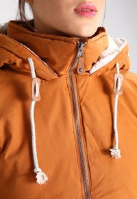 Orange jacka med hög krage, dragkedja och vita dragsnören. Har en fuskpälsfoder och läderdetaljer på dragsnörena.