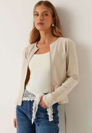 Ung kvinde iført en beige lynlåstrøje over en hvid top med blondedetaljer og blå jeans, stående op ad en lys væg.