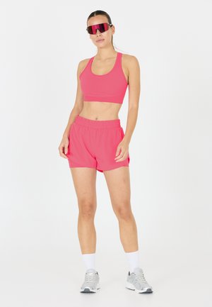 Roze atletische crop top en shorts van rekbaar materiaal, met een ontspannen pasvorm. Accessoires omvatten een paar oversized rode zonnebrillen en grijze sneakers.