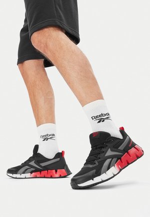 Czarne buty sportowe z czerwonymi akcentami, wykonane z siateczki i materiału syntetycznego, z teksturowaną podeszwą i kontrastową, białą środkową podeszwą. Połączone z białymi skarpetkami z logo.