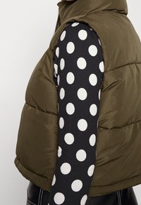 Olivgrön pufferväst med hög krage, över en svart långärmad topp med stora vita polka prickar mot en ribbad struktur.