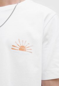 Witte katoenen t-shirt met een subtiele oranje zonprint met stralen. Korte mouwen, ronde hals en een textuurafwerking.