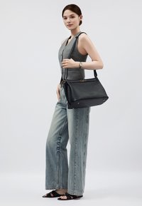 Borsa a tracolla in pelle nera opaca con dettagli dorati, dotata di chiusura con zip e tracolla regolabile. Modello indossa una maglietta grigia e jeans a gamba larga.