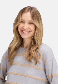 Junge Frau mit langem blonden Haar, die lächelt, trägt einen grauen Pullover mit beigen horizontalen Streifen, vor einem einfarbigen hellen Hintergrund.
