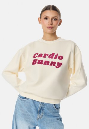 Młoda kobieta o gładkich włosach nosi kremową bluzę z wyraźnym różowym napisem "cardio Bunny" oraz jasnoniebieskie jeansy, stojąc z rękami na biodrach.