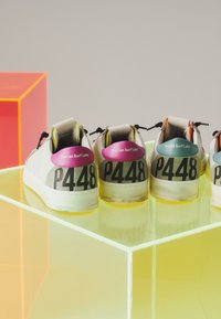 Quatre baskets blanches avec des languettes de talon colorées et "P448" imprimé au dos, exposées sur une surface translucide éclairée en jaune.