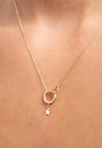 Gouden ketting met een cirkelvormige hanger en een kleine stervormige bedel, weergegeven tegen een huidskleurige achtergrond. De ketting is fijn en delicaat.