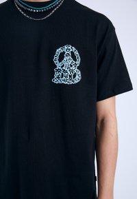 Camiseta de algodón negra con un gráfico de patrón floral en azul claro en el lado izquierdo del pecho. Diseño de cuello redondo con mangas cortas.
