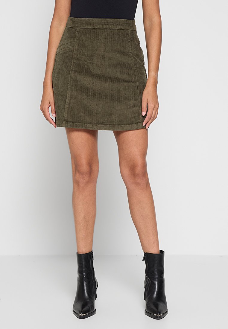 Olijfgroene corduroy minirok met opvallende naaddetails, hoge taille en gestructureerde vorm, gecombineerd met zwarte enkellaarsjes.