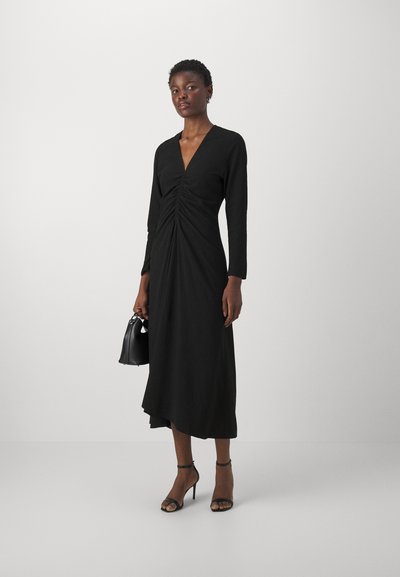 Vestido midi negro de manga larga, con un escote en V profundo y detalles fruncidos en la parte delantera, combinado con unos zapatos de tacón negros con tiras y un bolso.