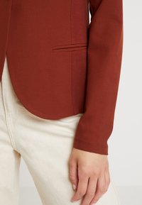Blazer couleur rouille en tissu lisse, avec un ourlet arrondi, une poche latérale et des manches ajustées, associé à un pantalon texturé crème.