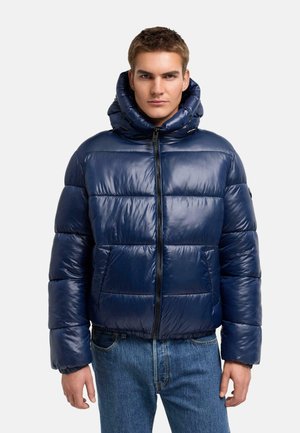 Homme portant une doudoune bleu marine avec une capuche et une fermeture éclair à l’avant, associée à un jean bleu, debout devant un fond uni.